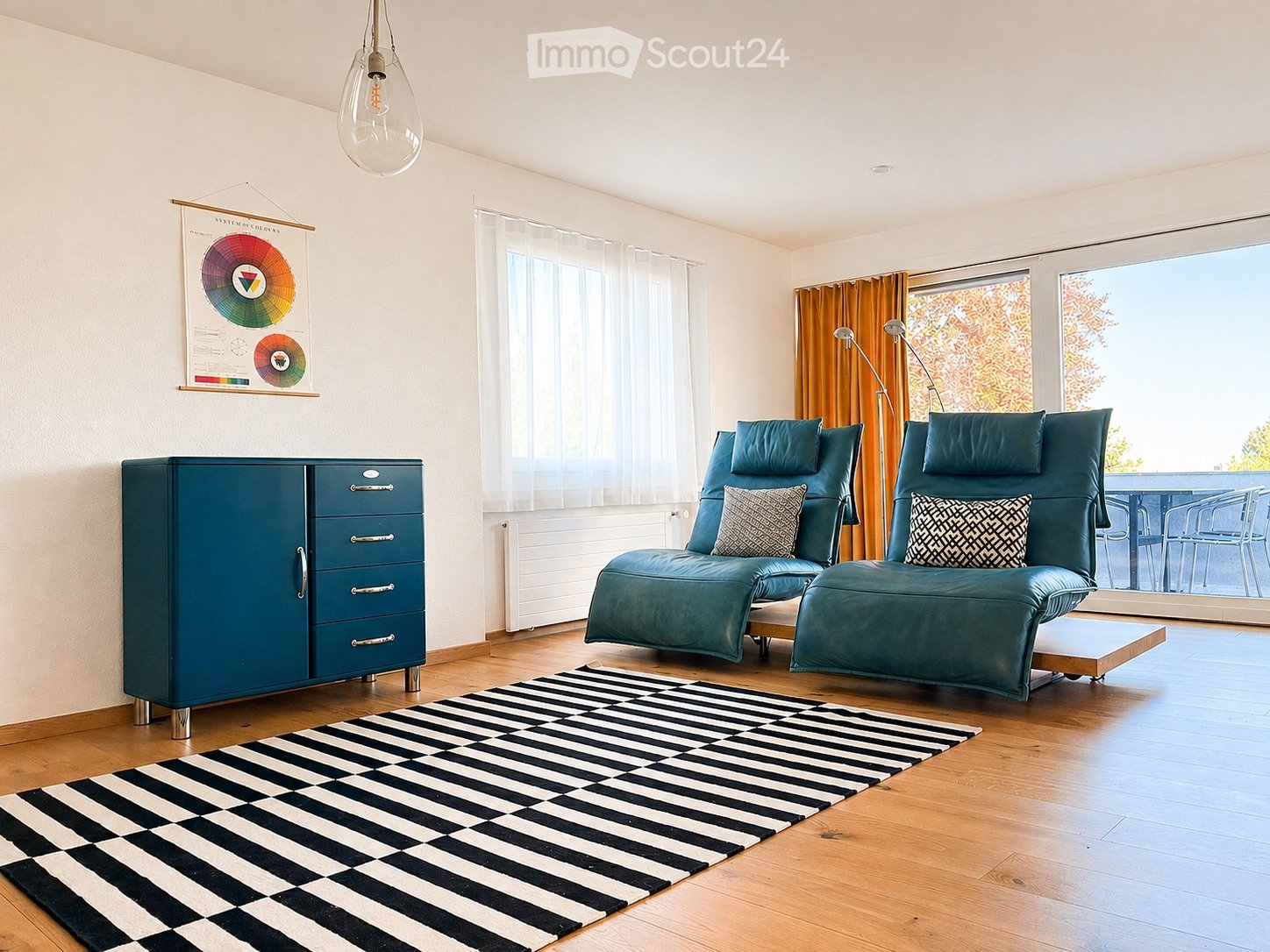 Modern living room with wooden flooring, large windows, teal sofa and chairs, black and white rug, cabinet, floor lamp, and a colorful poster on the wall.
