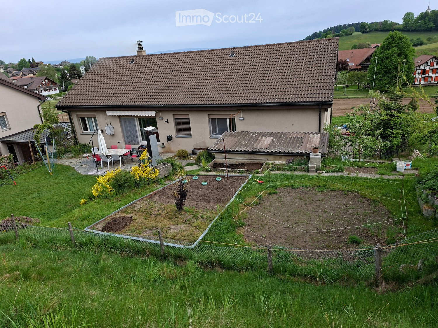 A well-maintained garden with a lawn, vegetable beds, a small patio area with outdoor furniture, and a house with a tiled roof in the background.