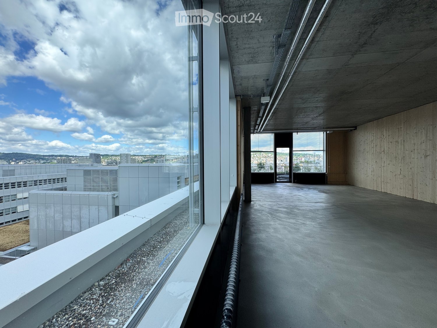 Modern balcony with large glass windows, concrete floor, and a view of the cityscape and surrounding buildings.