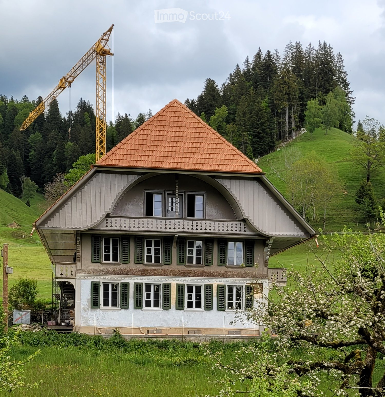 A two-story house with a red-tiled roof, white walls, and green shutters, surrounded by a well-maintained lawn and trees. A crane is visible in the foreground, indicating construction or renovation.