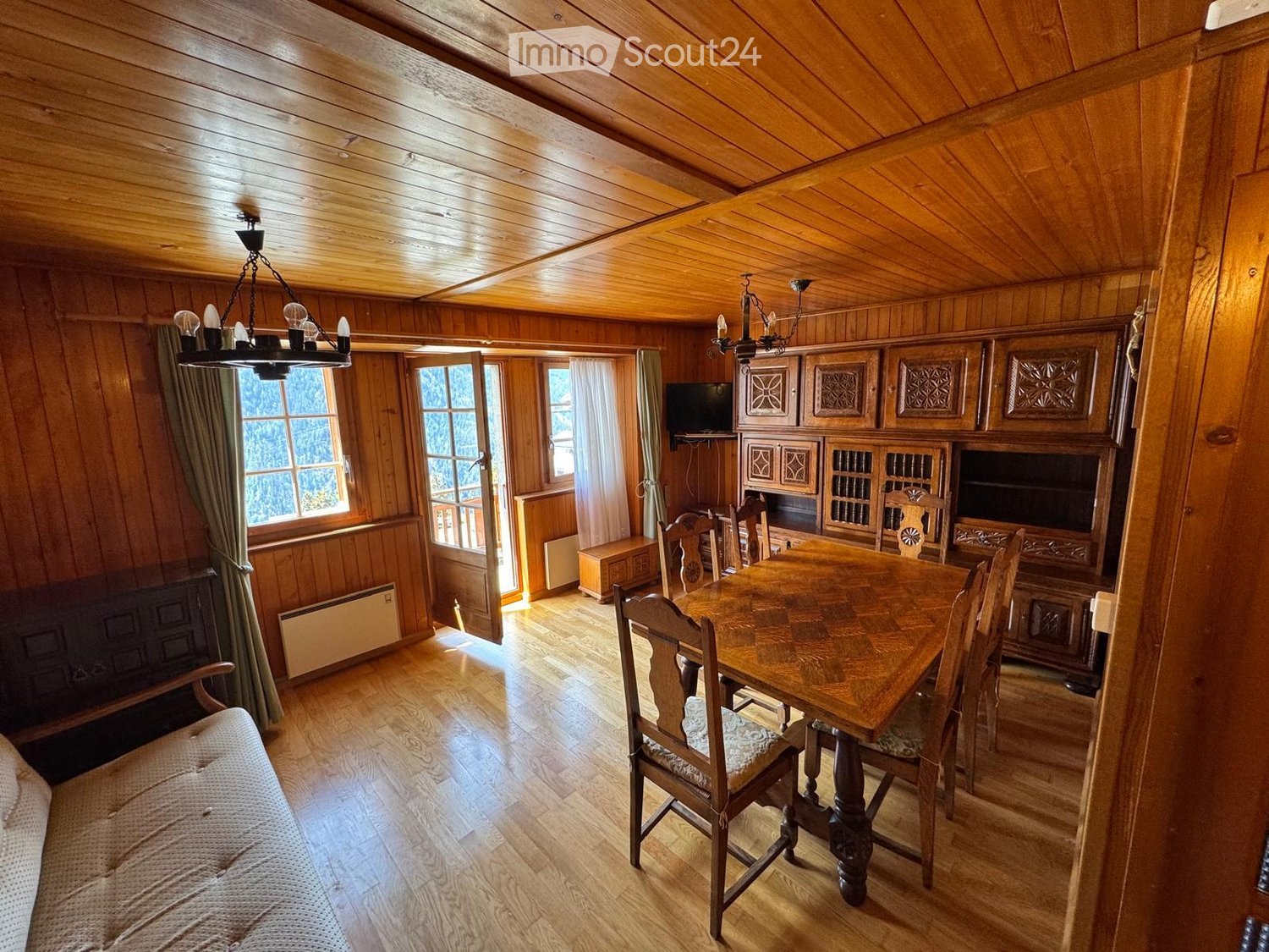 Cozy wooden-paneled room with a dining area, sofa, and cabinetry, featuring large windows and a chandelier.