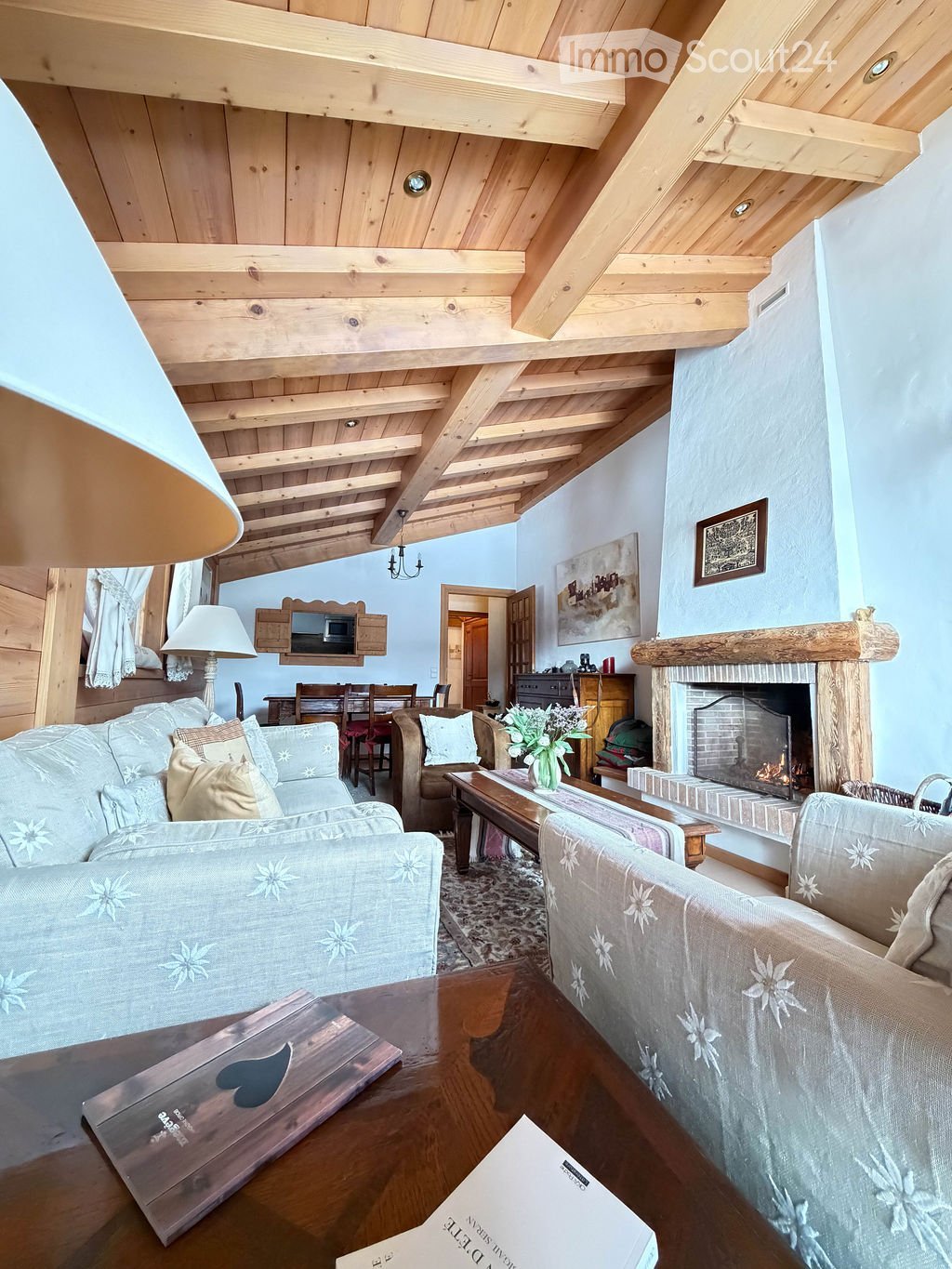 Cozy living room with exposed wooden beams, a fireplace, a sofa, armchairs, coffee table, and dining area visible in the background.