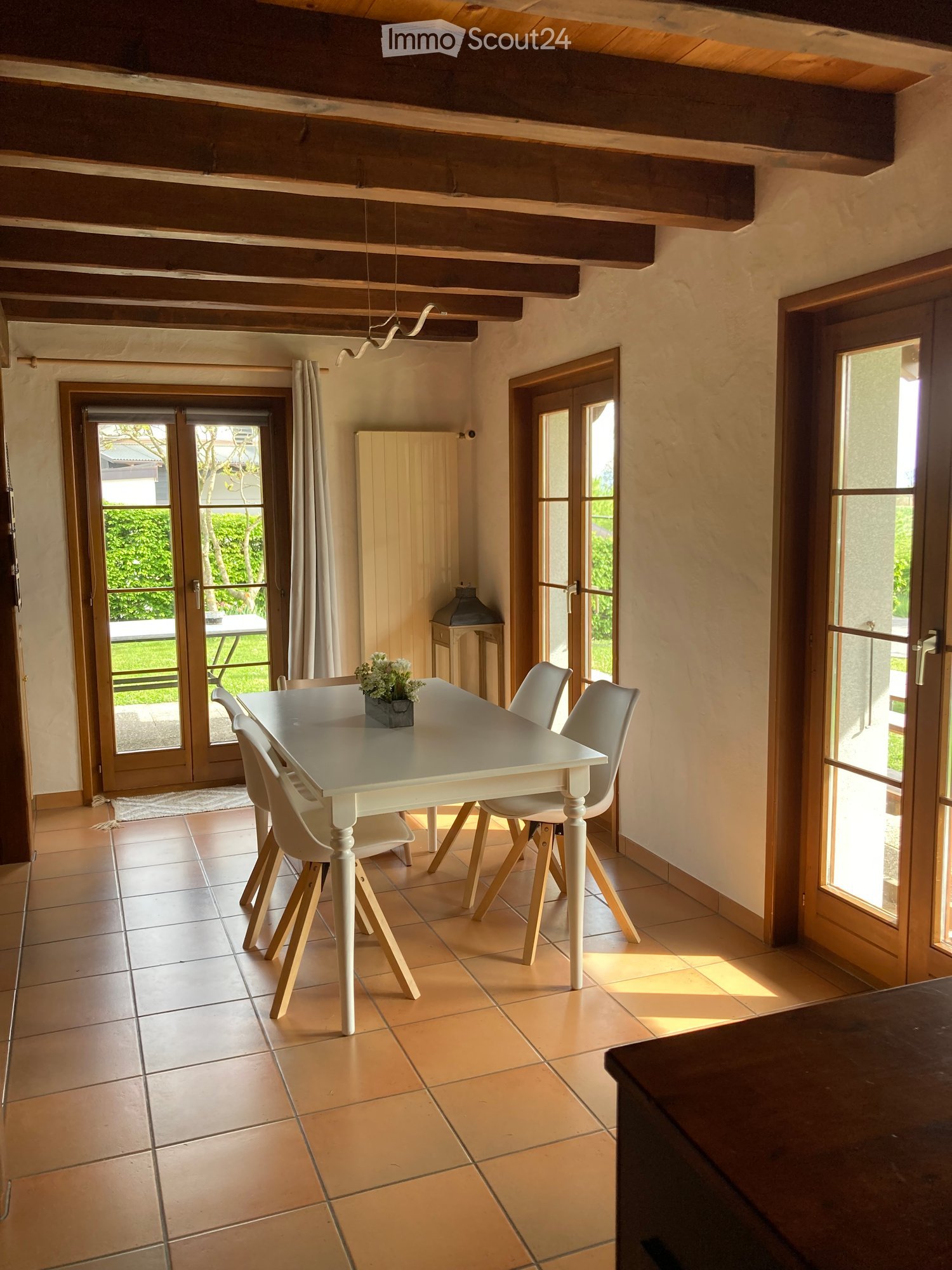 Bright dining area with a white dining table, four white chairs, a flower vase, and a wooden cabinet. Large windows and sliding doors lead to an outdoor area.