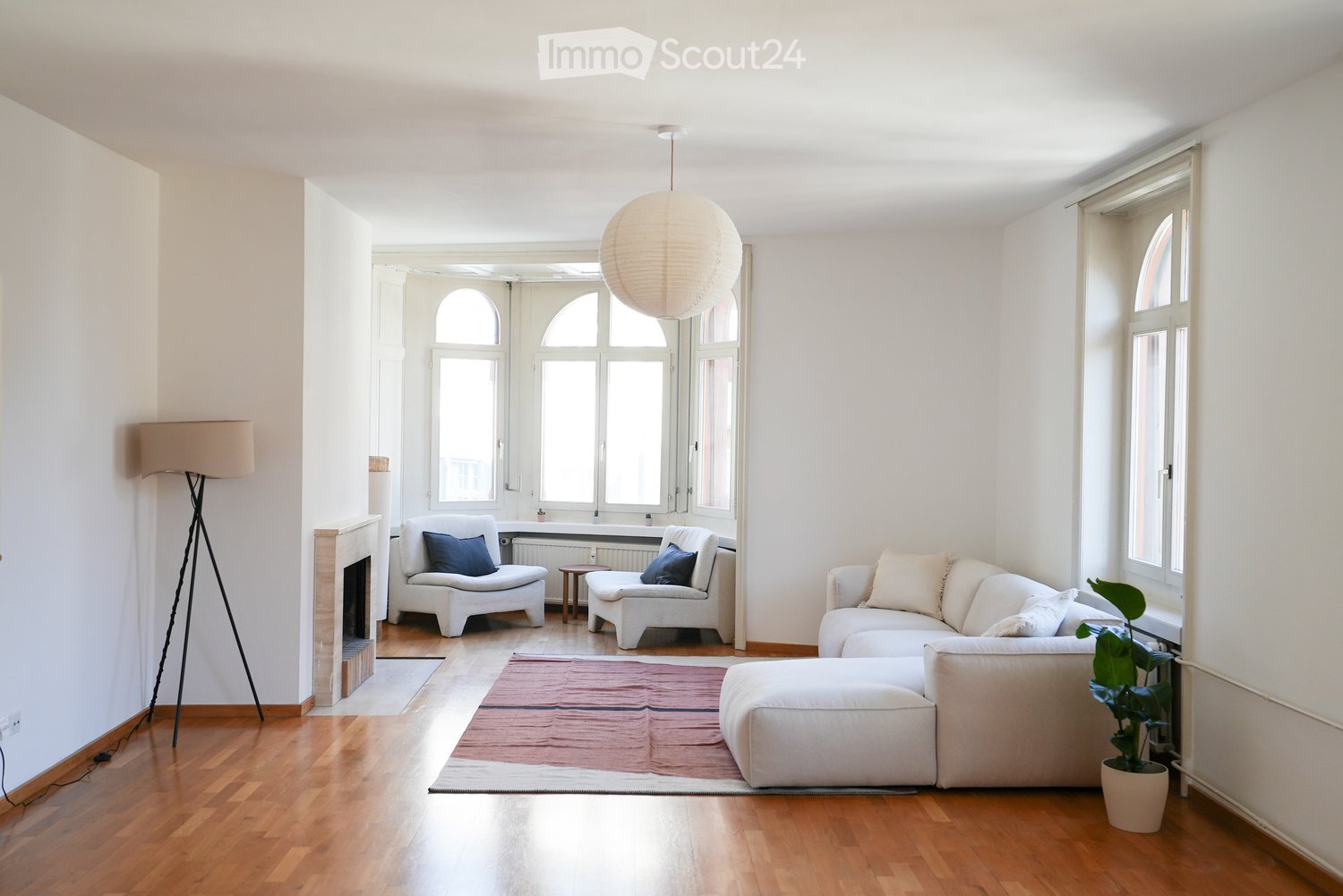 Bright living room with white walls, wooden flooring, large windows, a fireplace, a white sectional sofa, two white armchairs, a small wooden table, a floor lamp, a pendant lamp, and a potted plant.