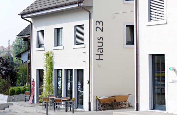 white exterior, large glass windows, 'Haus 23' written on the wall, exterior steps, garden, chairs, and tables in front of the house, brown trolley on the side