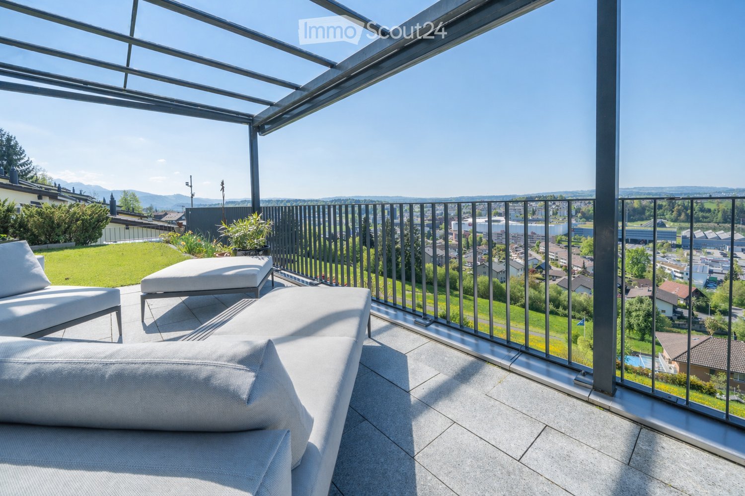 Modern balcony with glass roof and metal railing, featuring white outdoor furniture including a sofa and chairs, overlooking a cityscape with greenery and distant mountains.