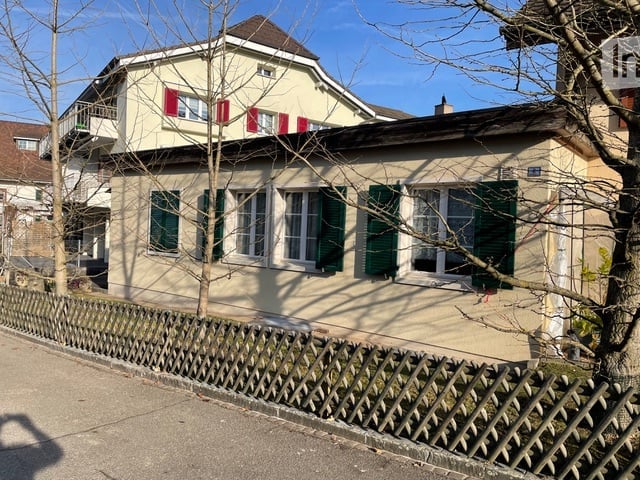 House. Two-story house with cream-colored walls, red shutters on the upper level, green shutters on the lower level, and a wooden fence in front.