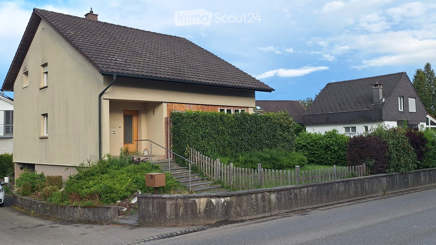 Two-story house with beige siding, dark roof, and a small front porch with steps and railing. The house is surrounded by a well-maintained garden with hedges and a wooden fence.