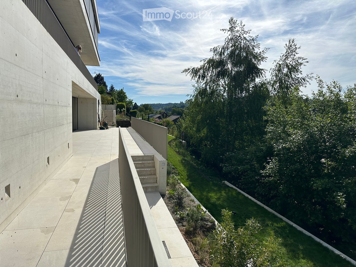 Modern concrete balcony with steps leading down to a landscaped garden area, surrounded by tall trees and greenery.