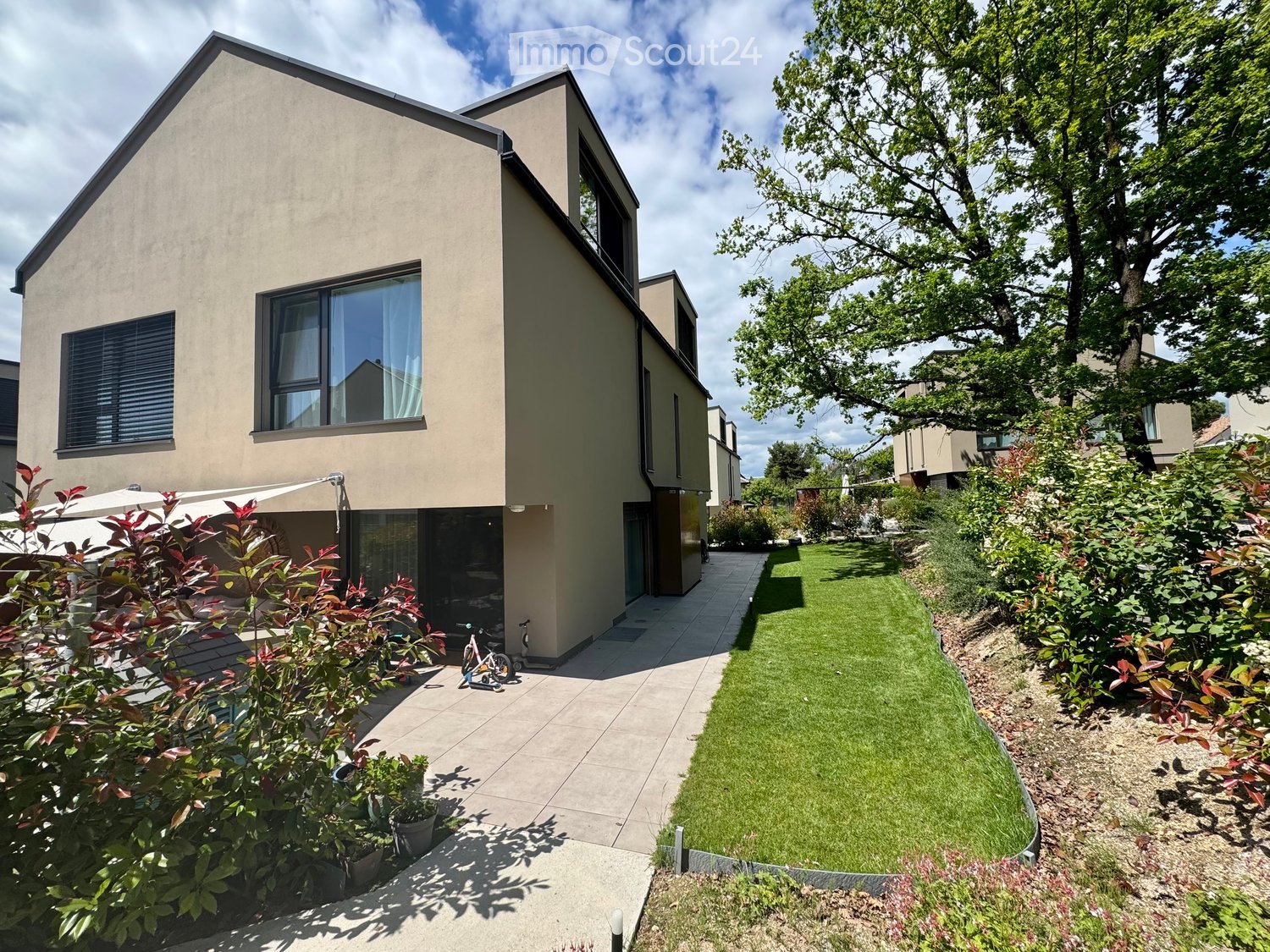 Modern two-story house with beige exterior, large windows, and a well-maintained garden with a lawn, shrubs, and a pathway leading to the entrance.