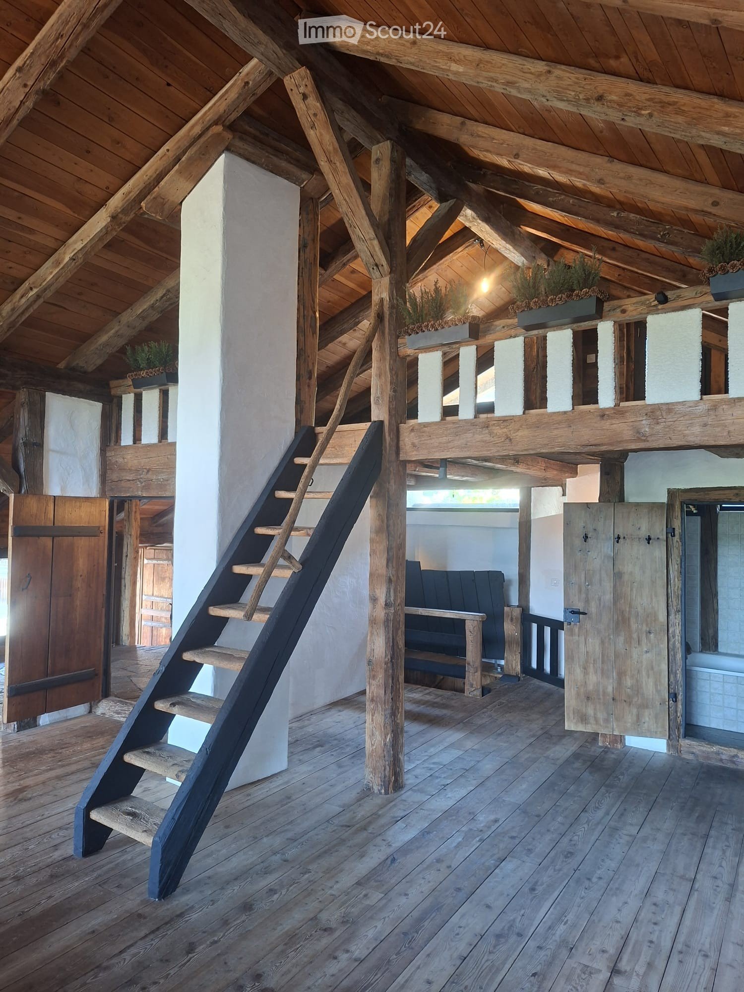 Loft-style room with wooden beams, exposed wooden floor, a wooden staircase leading to an upper level, a sofa, and potted plants on the upper level.