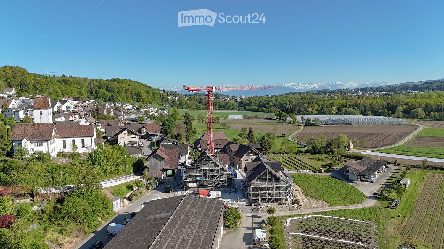 Aerial view of a rural village with traditional houses, a church, and a construction site with a crane in the background, surrounded by green fields and mountains.