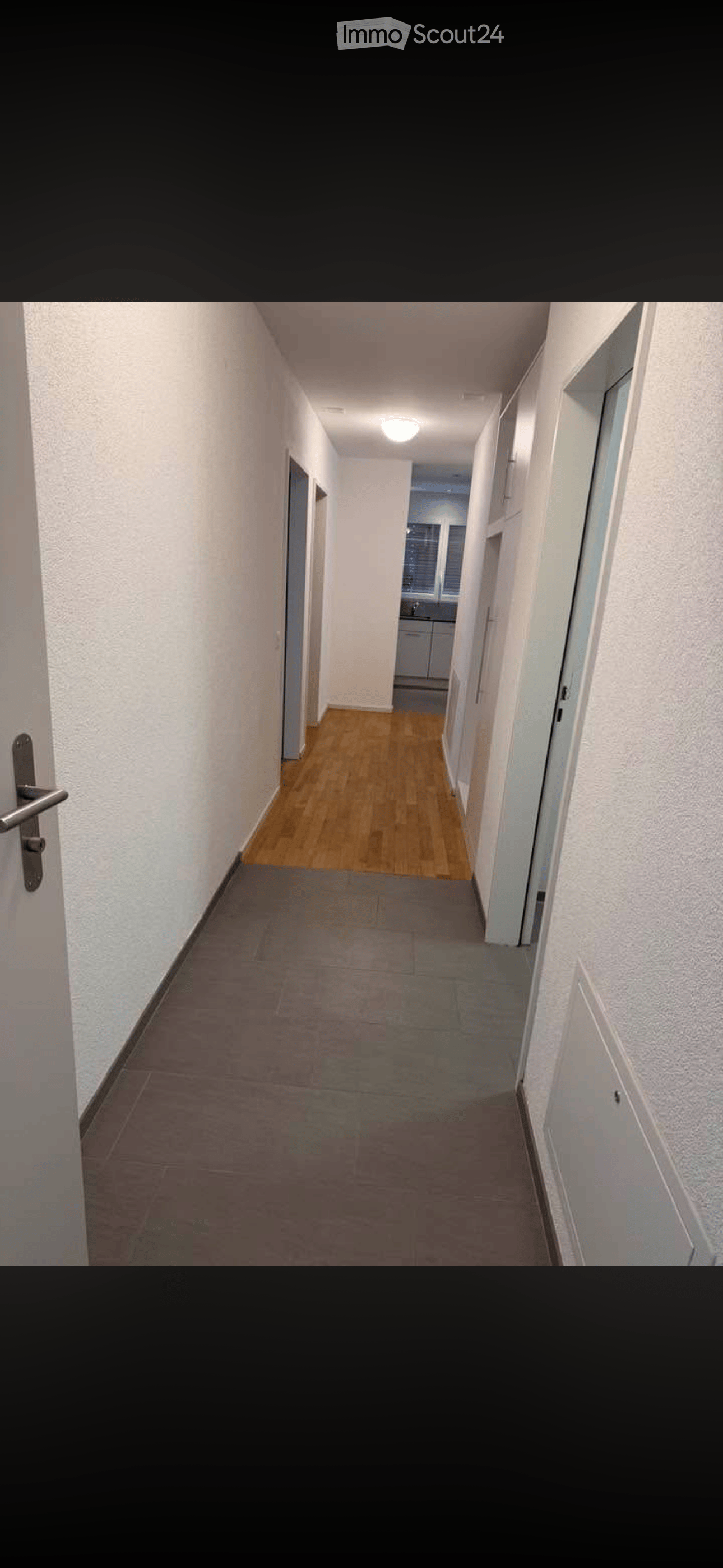 A long, narrow hallway with light-colored walls, wooden flooring in the center, and tiled flooring on the sides. Doors line both sides, and a small room with cabinets and a window is visible at the end.