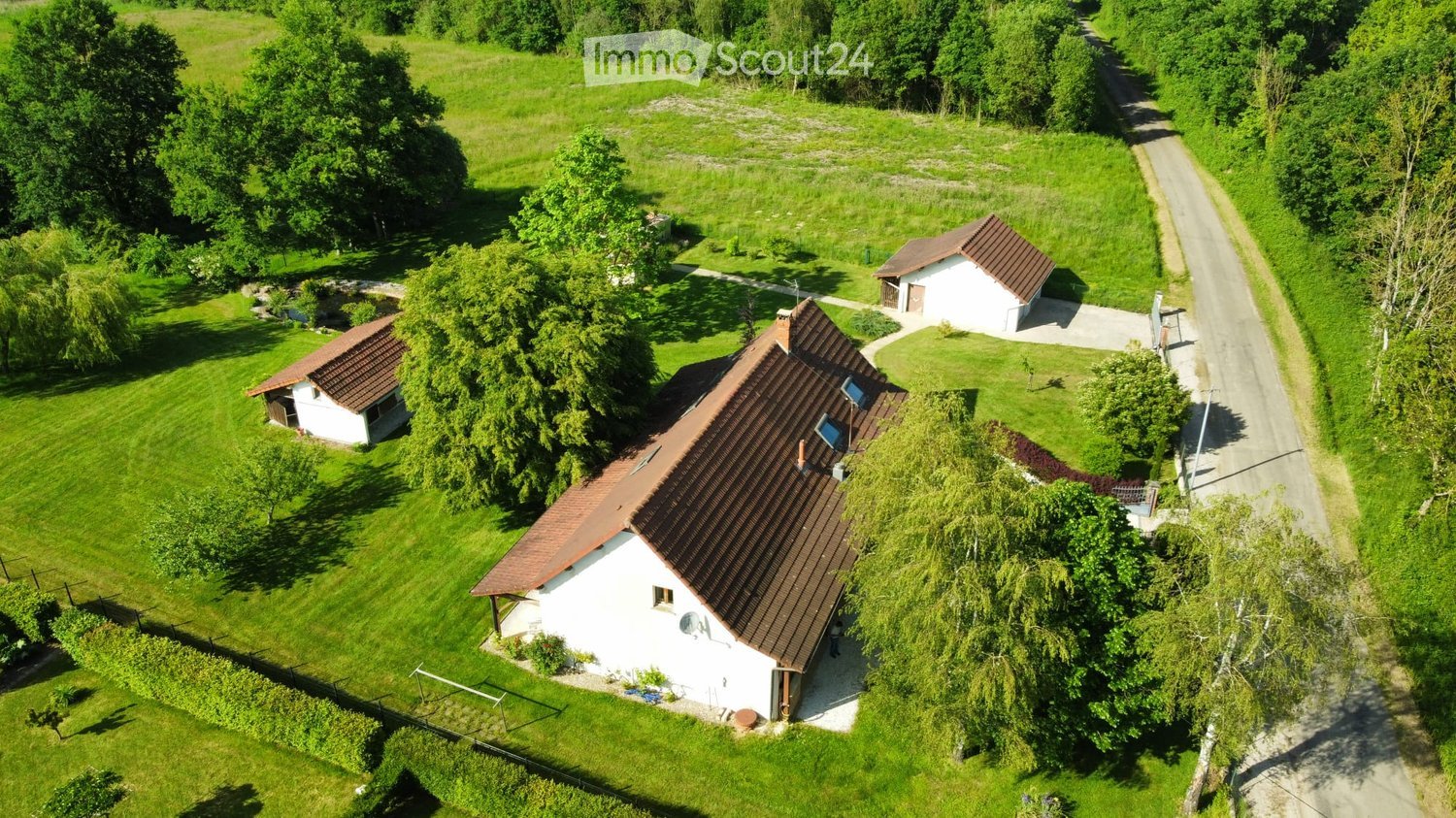 Aerial view of a single-story house with a brown tiled roof, surrounded by a well-maintained lawn and trees. The house has multiple windows and a small outbuilding nearby.