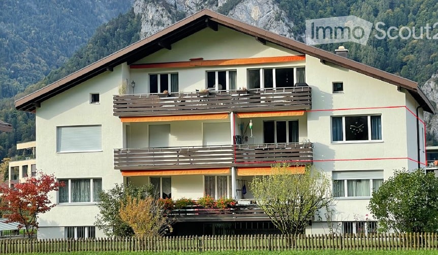 Two-story house with balconies, surrounded by greenery and mountains.
