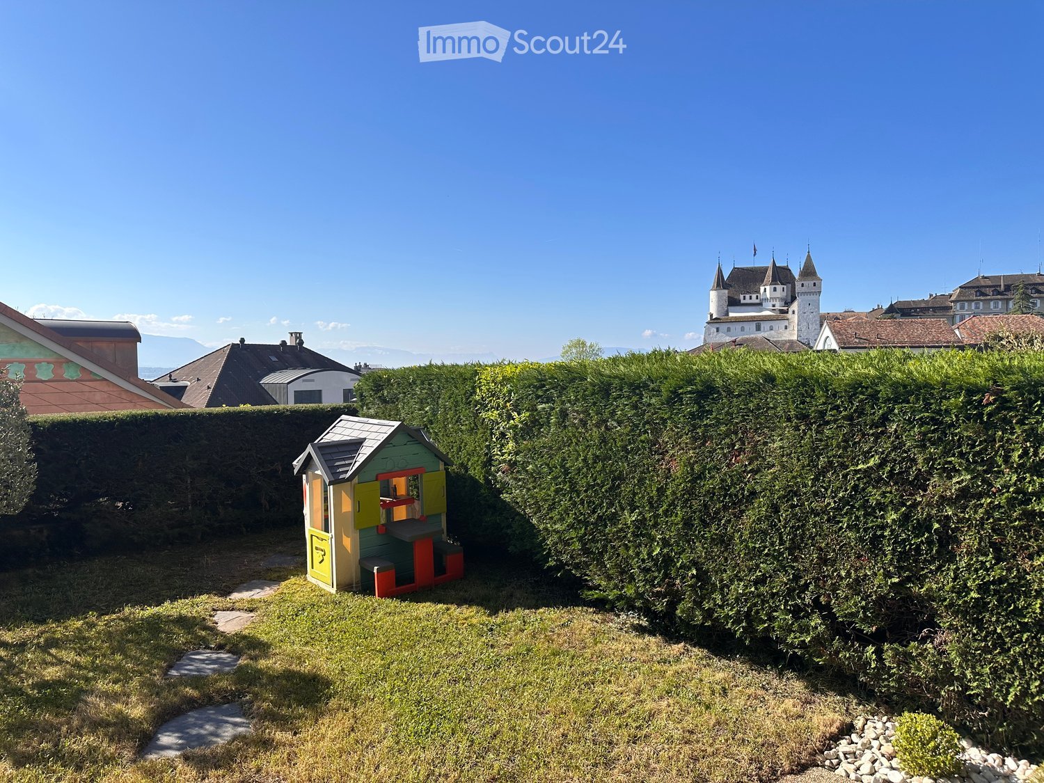 A small playhouse in the middle of a well-maintained garden with a hedge, surrounded by a stone pathway.