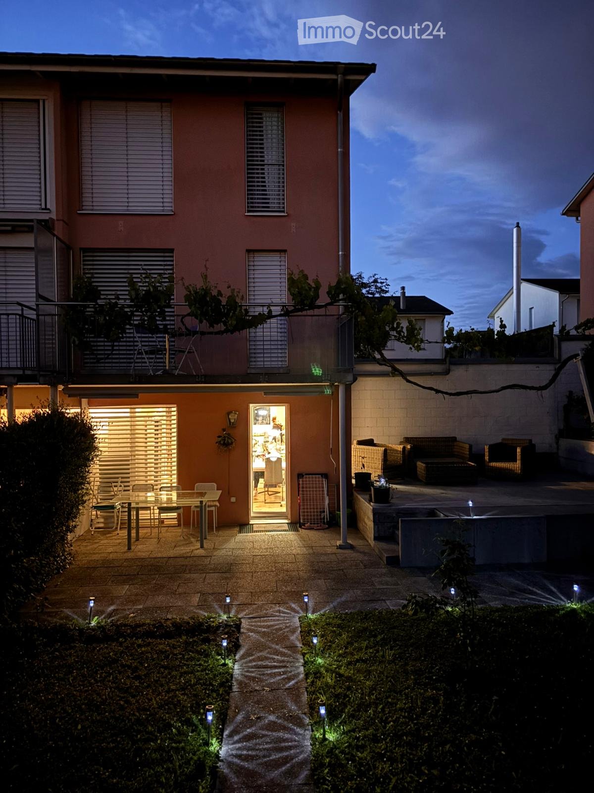 Evening view of a well-lit garden area with a dining table and chairs, a sofa and ottoman, and decorative lighting along a pathway.