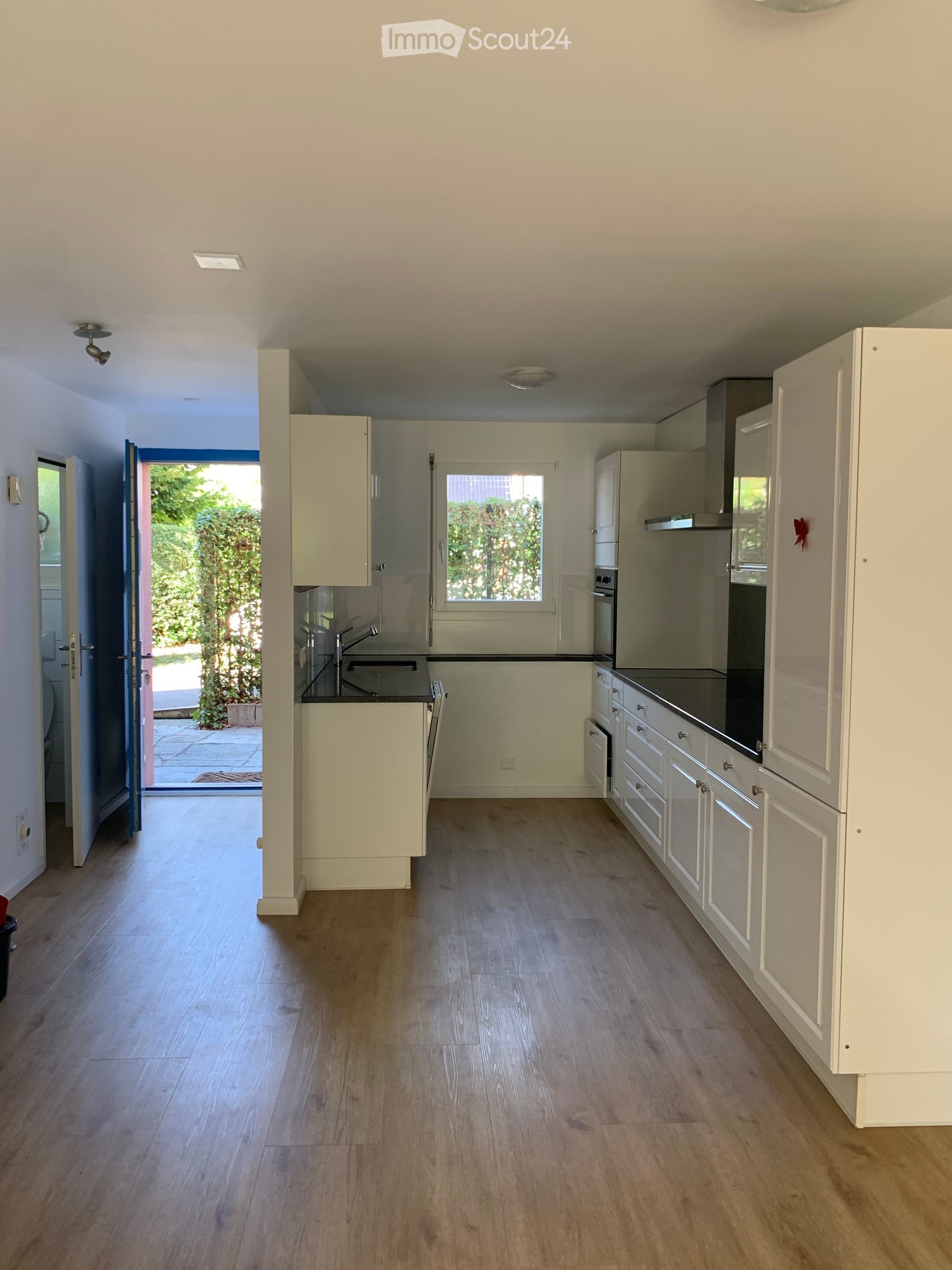 Modern kitchen with white cabinets, wooden flooring, and stainless steel appliances including a sink, oven, and refrigerator. A window provides natural light, and a door opens to an outdoor area.