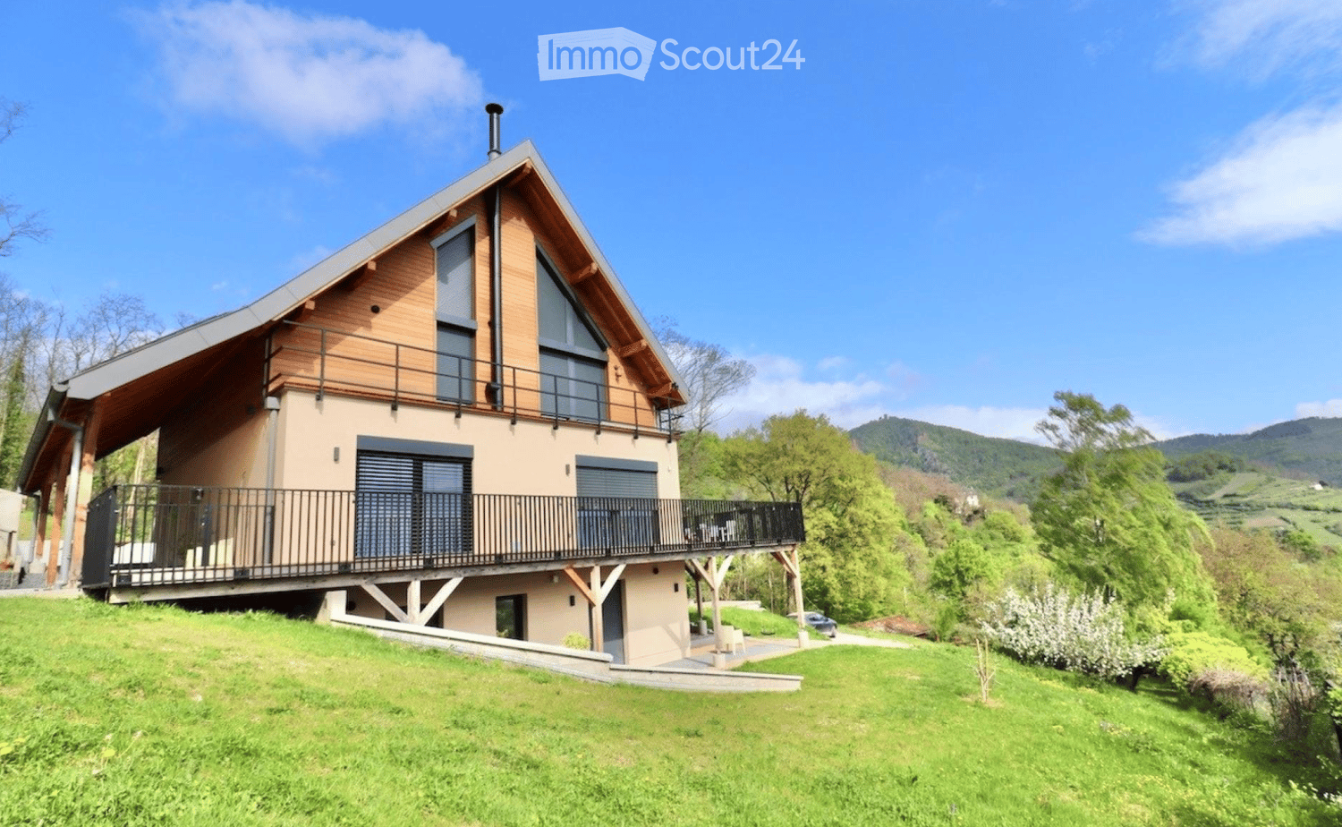 House. Two-story house with a modern design, featuring a wooden facade, large windows, and a spacious balcony overlooking a green, hilly landscape.