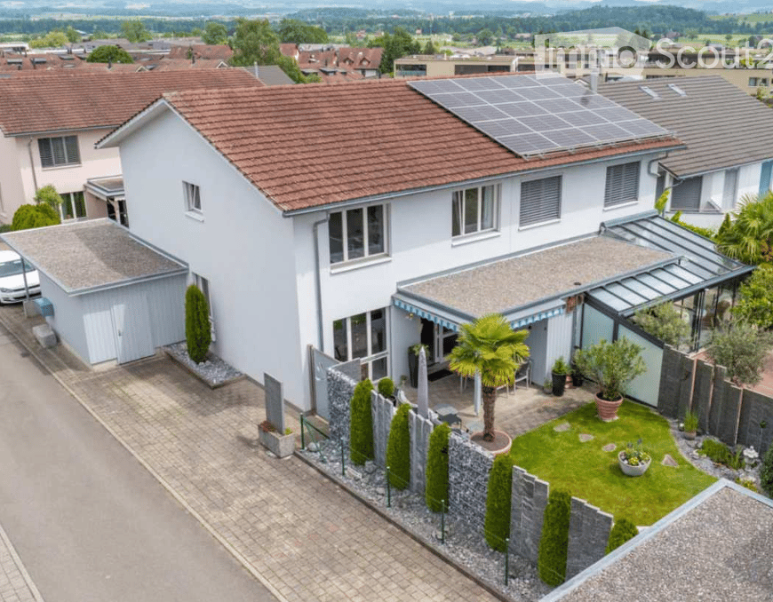 House. A two-story white house with a red-tiled roof, solar panels on the roof, and a modern garage. The house features large windows and a well-maintained garden with a palm tree, shrubs, and a small lawn.