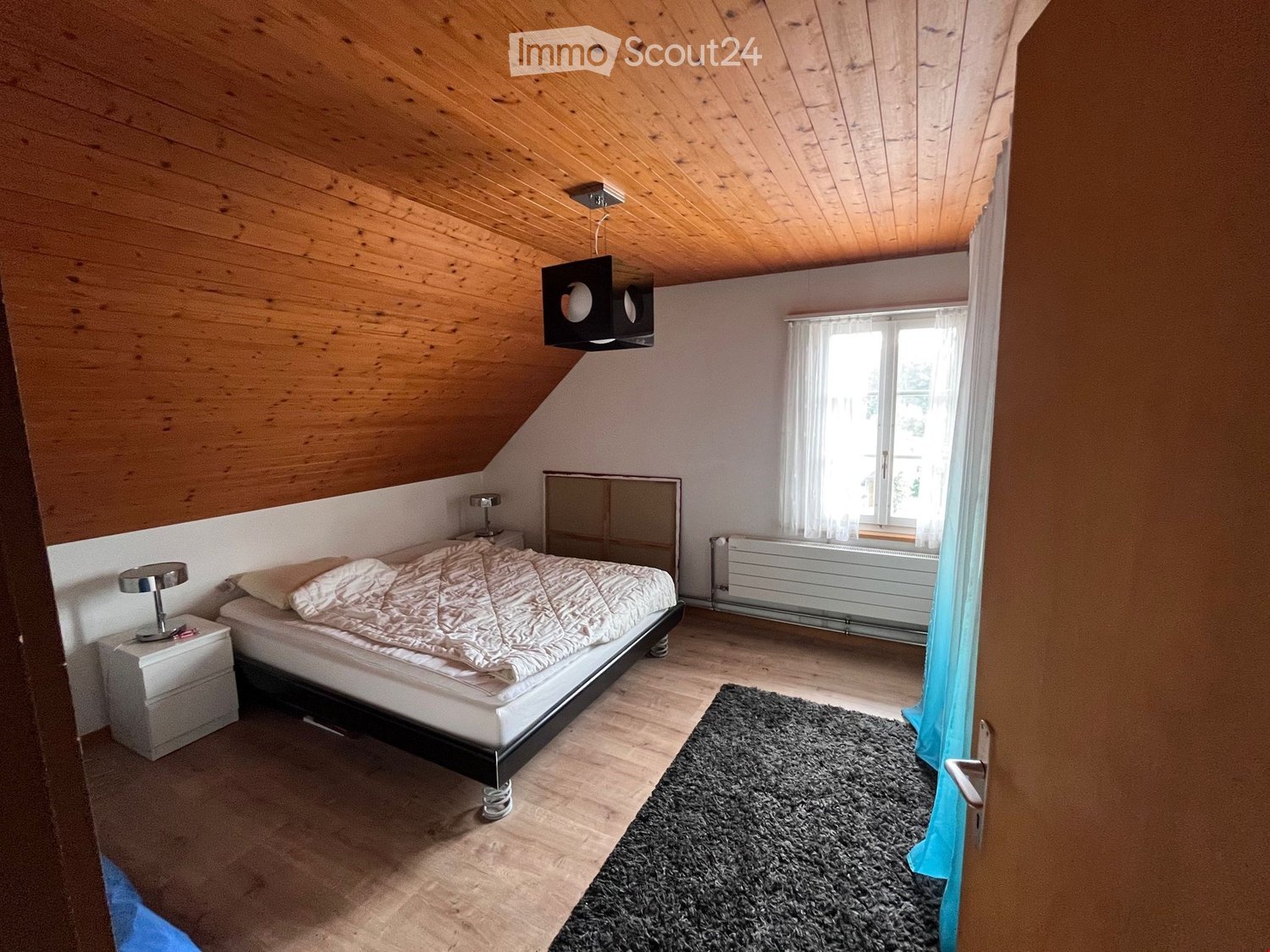 bedroom with bed, wooden ceiling, white walls, white nightstand, lamp, radiator, window with white curtains, rug