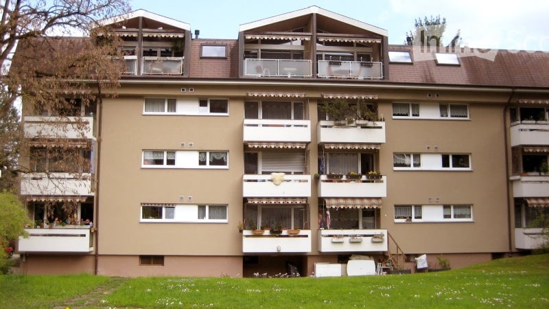 Multi-story residential building with multiple balconies and windows, surrounded by greenery.