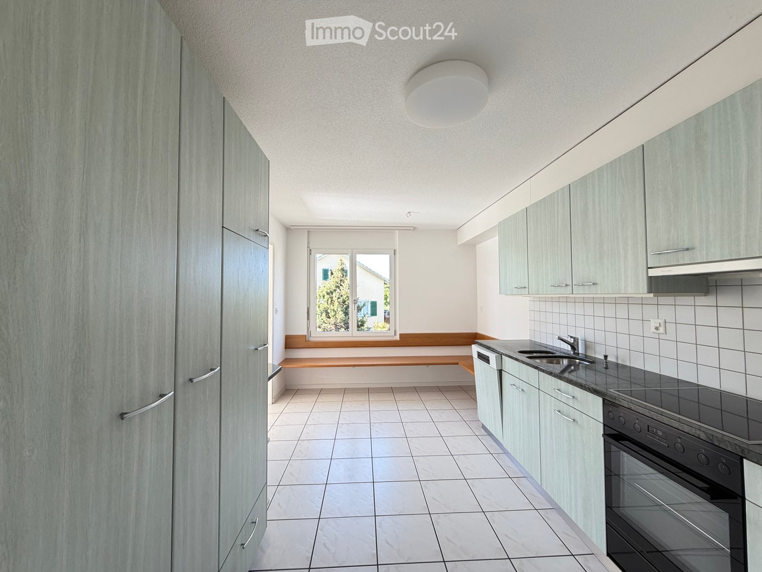 Modern kitchen with light grey cabinets, white tiled floor, and a window overlooking a green area. Features a sink, faucet, oven, and dishwasher.