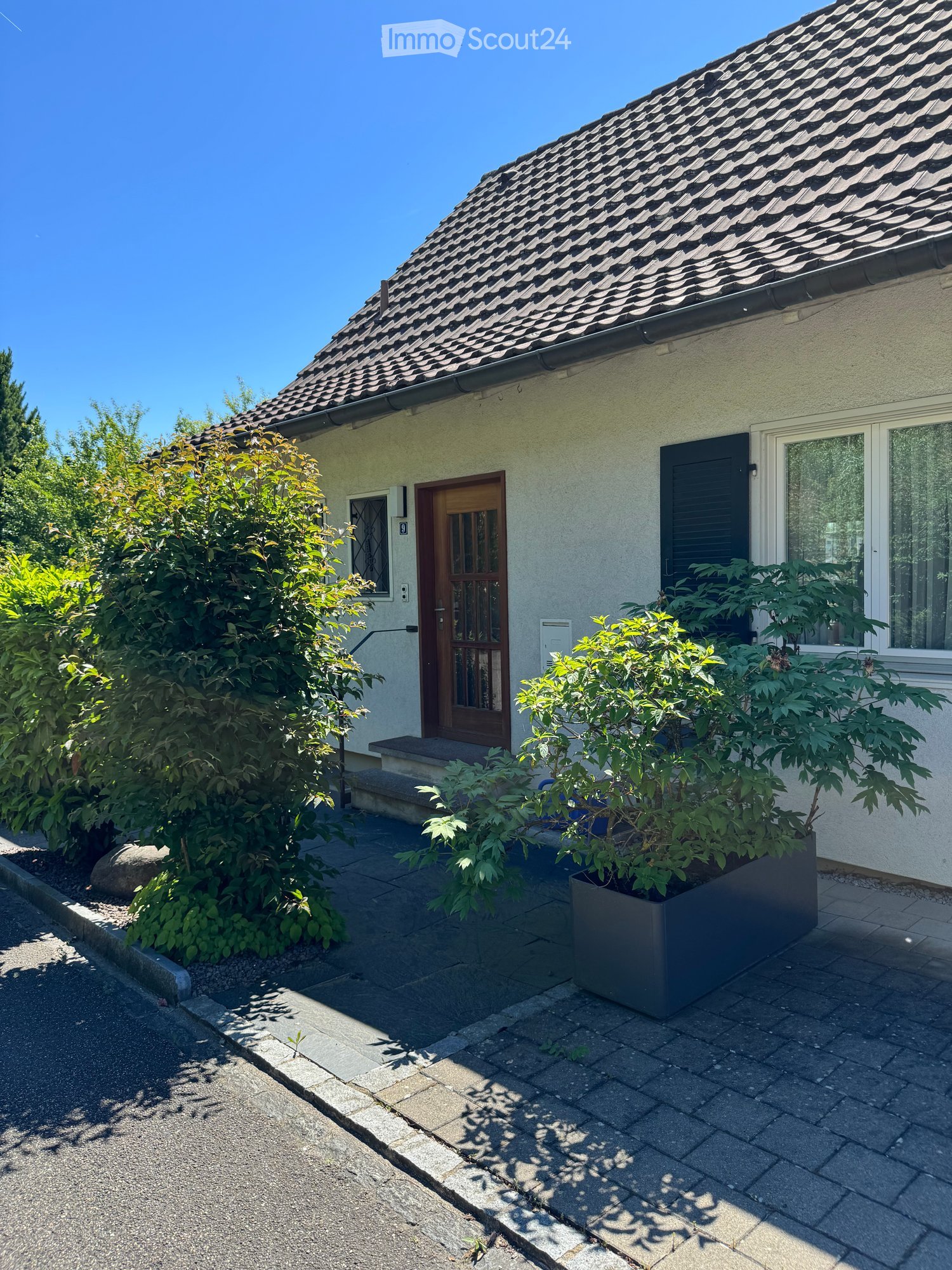 Single-story house with a tiled roof, light-colored exterior, and a wooden front door. The entrance has a small concrete step leading up to the door, surrounded by greenery and potted plants.