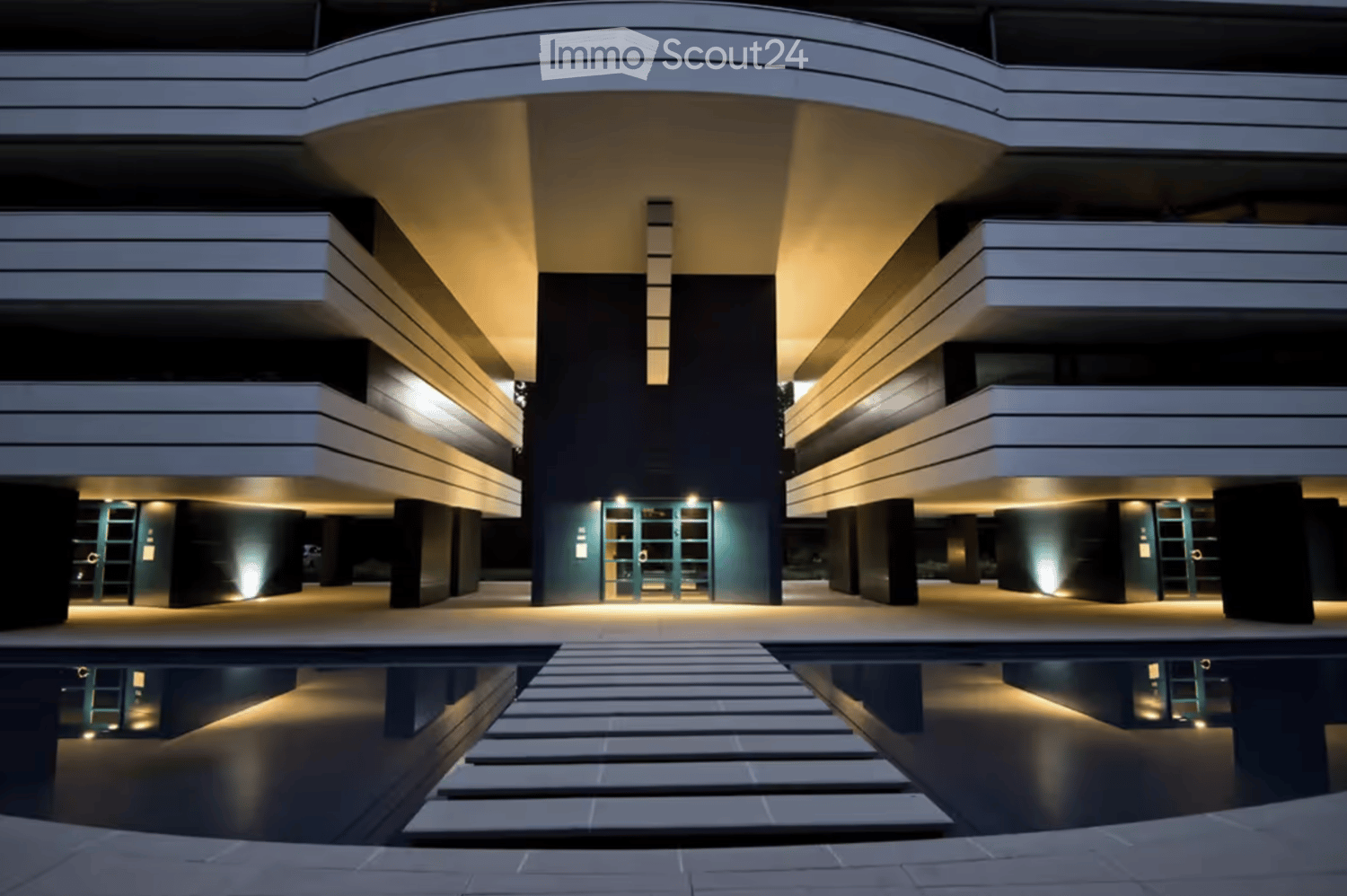Modern multi-story building with sleek, contemporary design featuring illuminated entrances and reflective water surface in front.