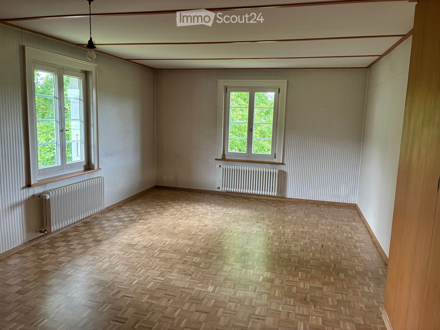 Empty room with wooden herringbone flooring, white walls, two windows, and radiators beneath each window.