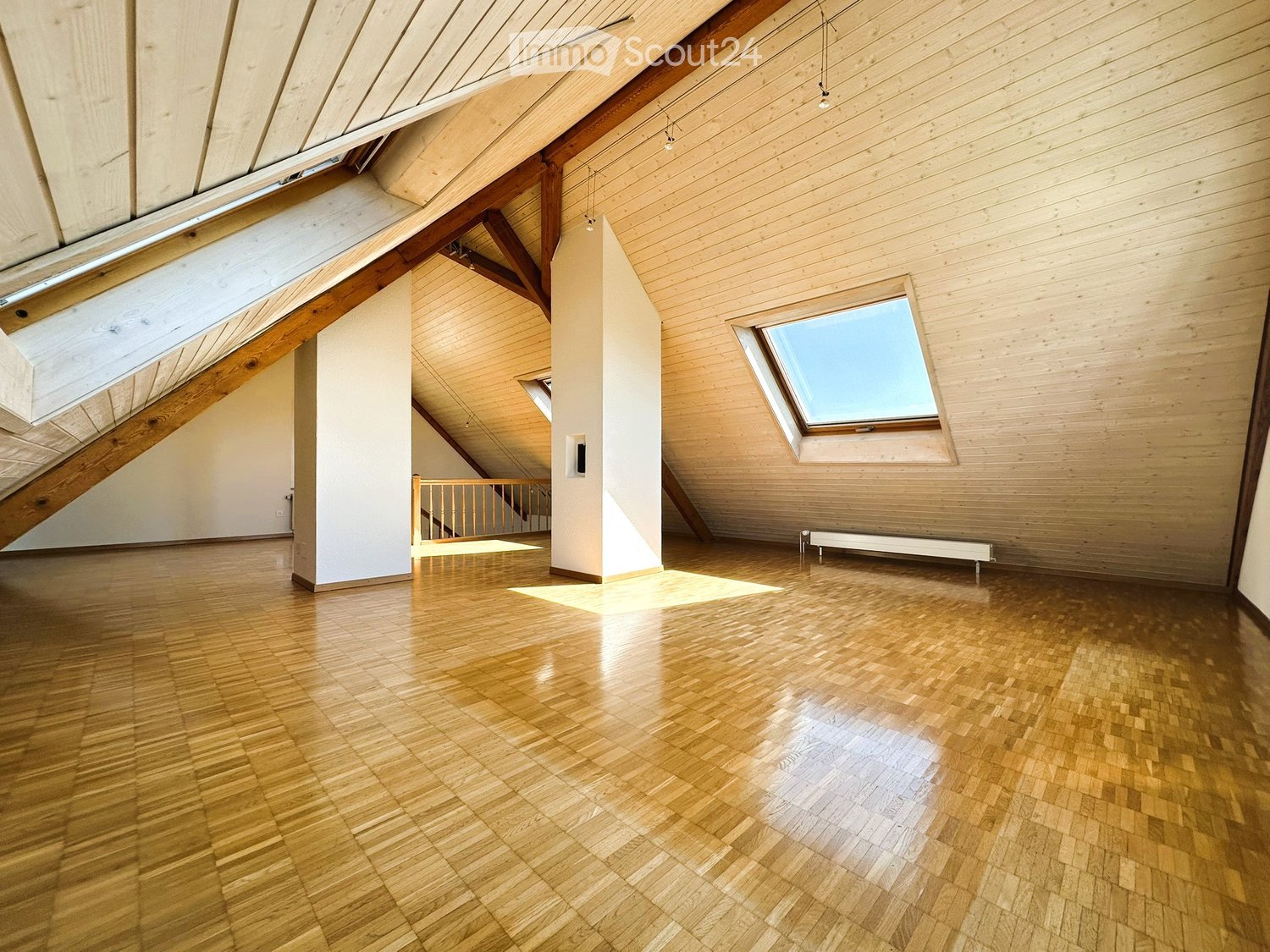 Bright, empty attic space with wooden floors, exposed wooden beams, skylights, and a small staircase leading to another area.
