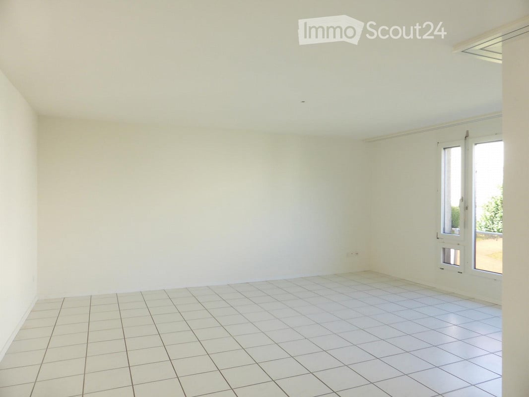 Empty room with white walls, tiled floor, and large windows allowing natural light.