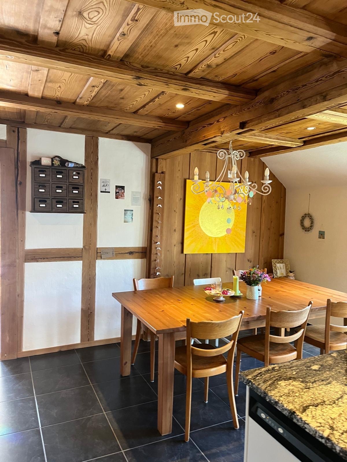 Cozy kitchen with wooden dining table, chairs, chandelier, and a colorful painting on the wall.
