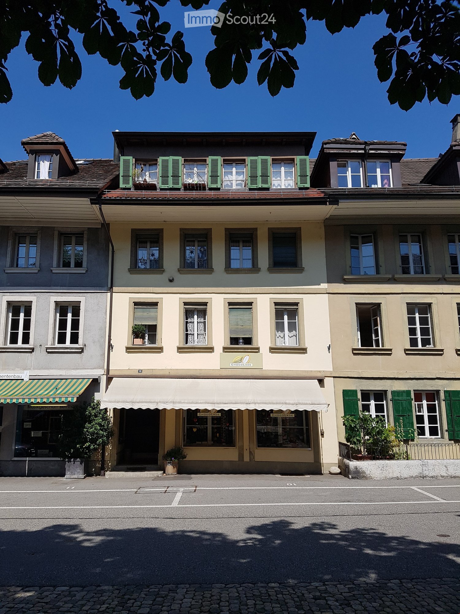 Three-story building, beige exterior, green shutters, awning over entrance, small plants at the entrance, clear sky