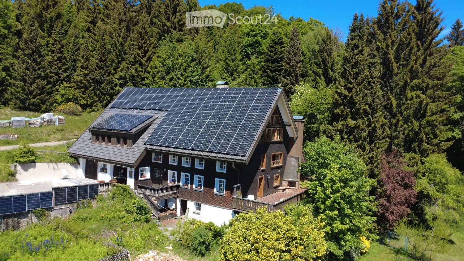 A two-story house with a dark brown exterior, white trim, and multiple windows. The roof is covered with solar panels, and the house is surrounded by lush greenery and trees.