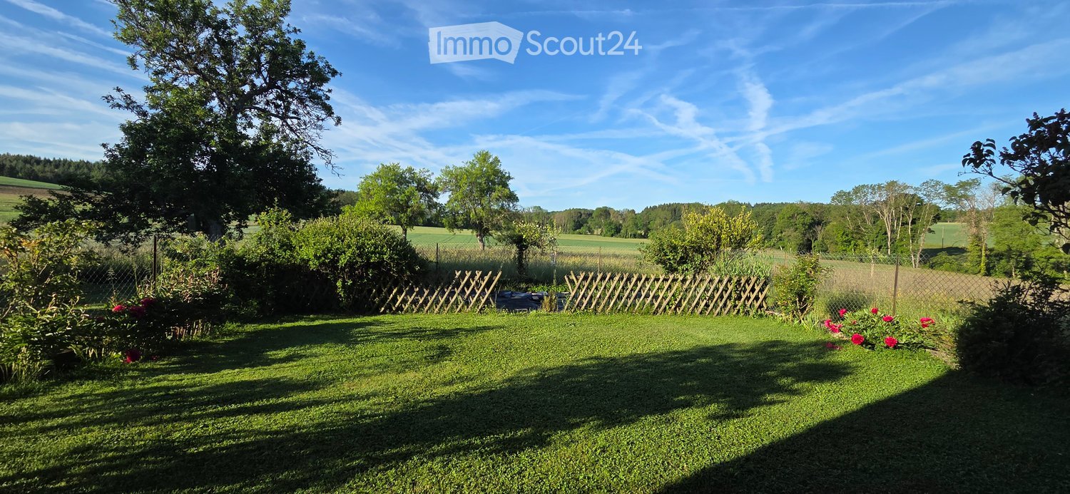 Well-maintained garden with a wooden fence, lush greenery, and a clear blue sky. Features include a neatly trimmed lawn, flowering shrubs, and a scenic rural backdrop with fields and trees.