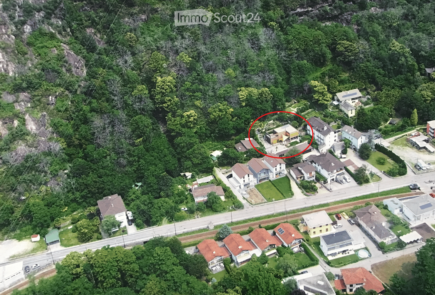 Aerial view of a residential area with multiple houses, surrounded by greenery and trees. The highlighted house is a modern two-story structure with a flat roof and large windows, situated on a hillside with a view of the surrounding landscape.