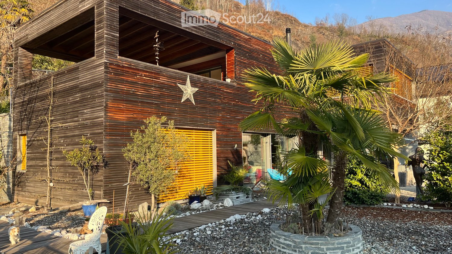 Modern wooden house with a contemporary design, surrounded by a landscaped garden with palm trees, potted plants, and a small seating area.