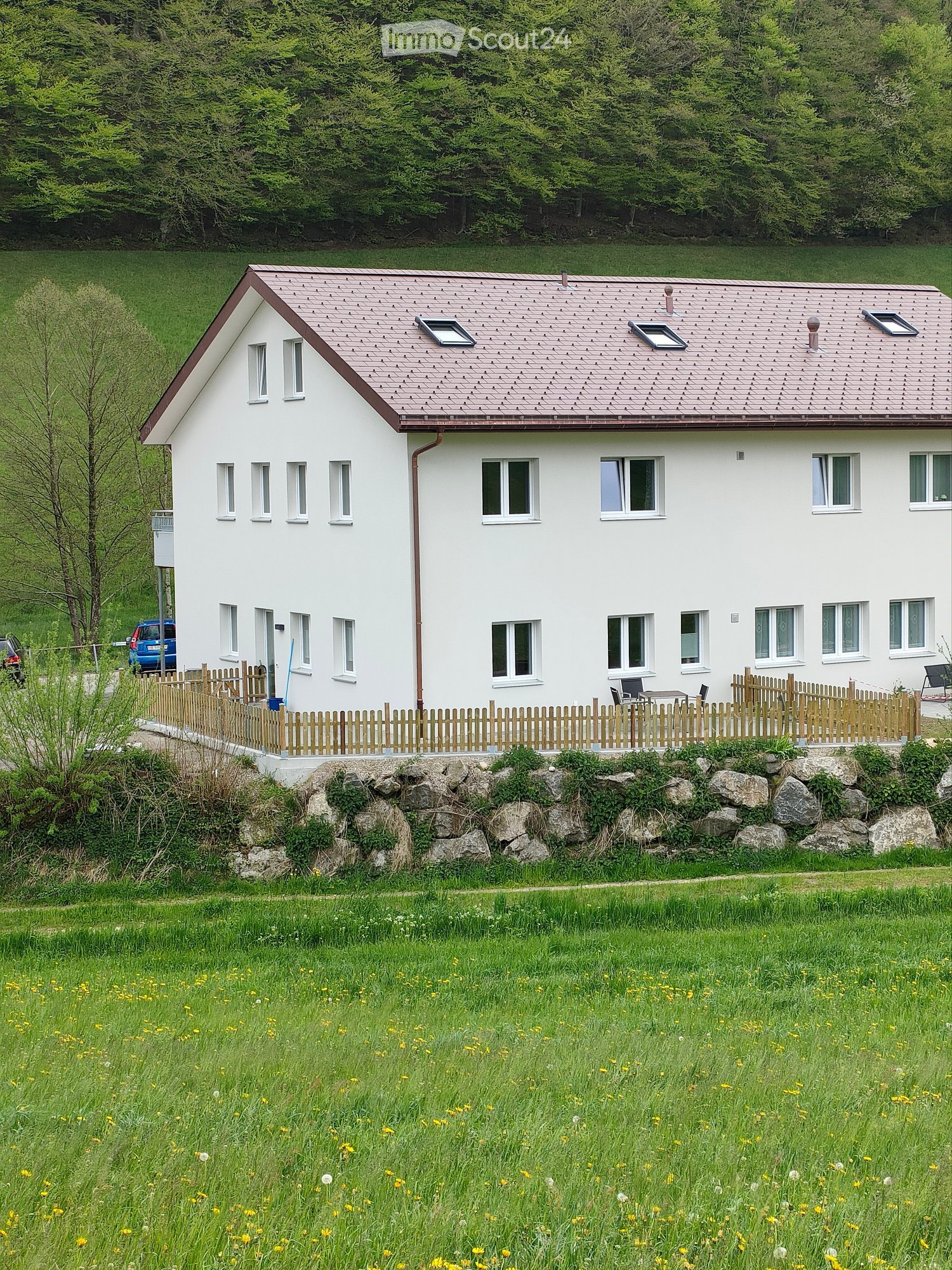 Single-story white house with a pitched roof, multiple windows, and a wooden fence surrounding the property. There is a small garden area with a table and chairs, and a stone retaining wall in front of the house.