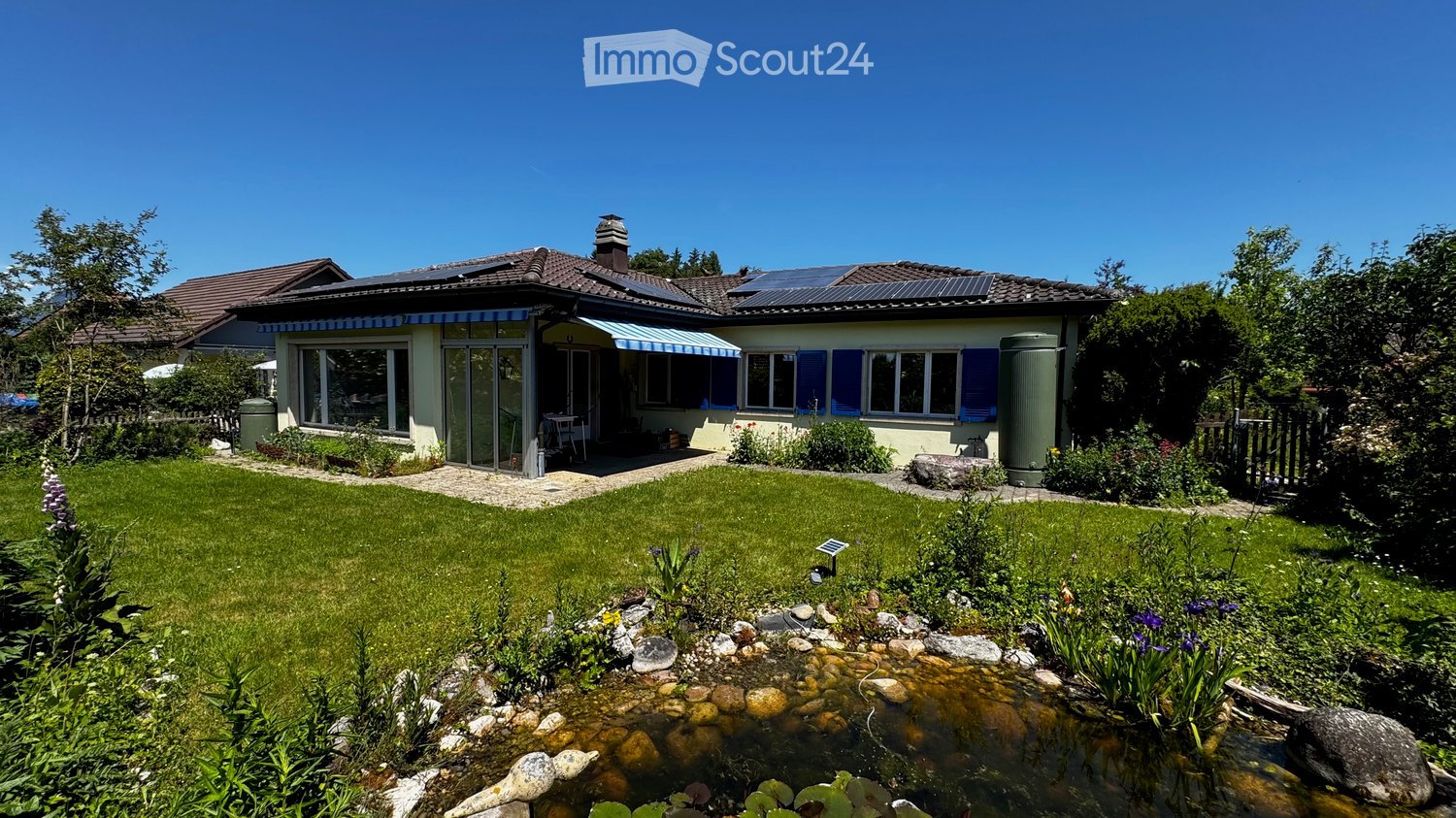 Two-story house with solar panels on the roof, blue shutters, and a well-maintained garden with a pond and patio area.