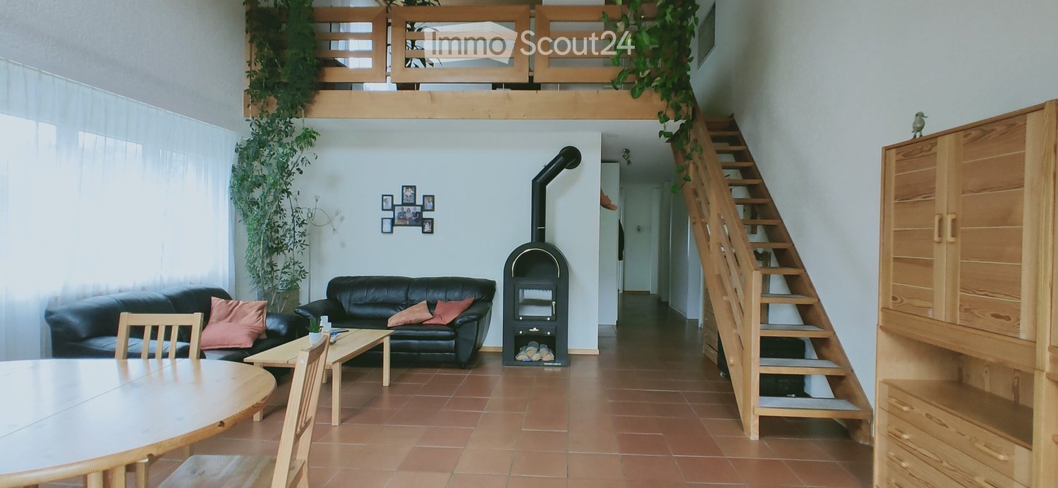 Living room with a black leather sofa, coffee table, dining table, chairs, and a wood-burning stove. There is a staircase leading to an upper level and a wooden cabinet on the right side.