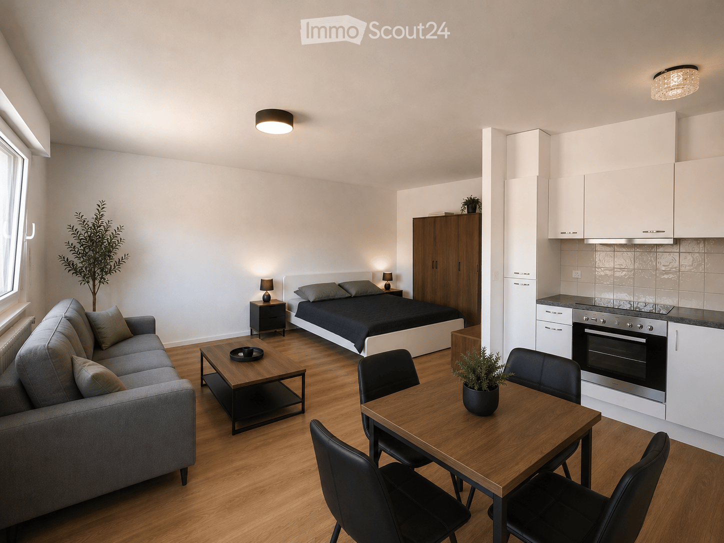 Modern, open-concept studio apartment with a grey sofa, coffee table, bed, nightstand, dining table, chairs, and kitchen area featuring white cabinets and an oven.