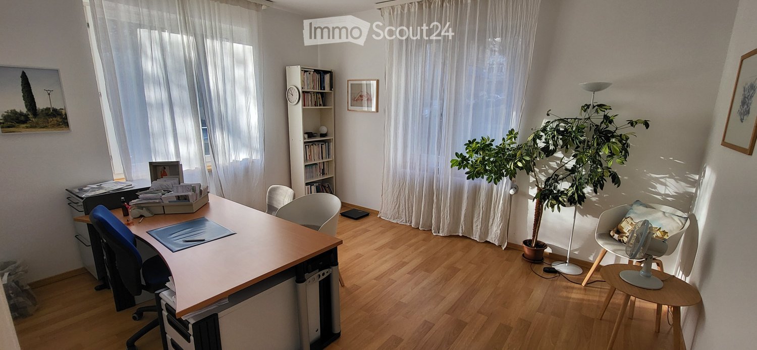 Brightly lit room with wooden flooring, large window with white curtains, desk with office supplies, bookshelf, potted plant, and a small fan on a stool.