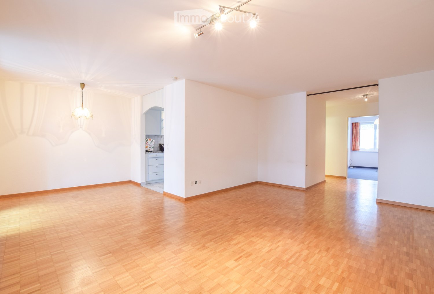 Empty room with wooden flooring, white walls, and a hanging light fixture. Adjacent kitchen area visible with cabinets and a countertop.