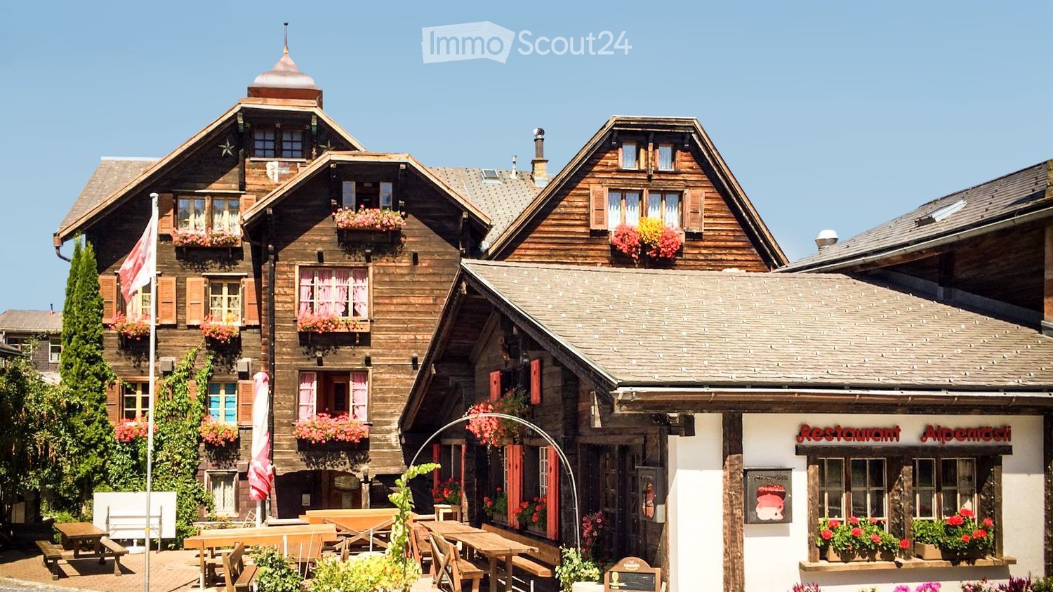 Traditional wooden chalet-style buildings with flower boxes, outdoor seating area, and a restaurant sign reading "Restaurant Alpenrose".