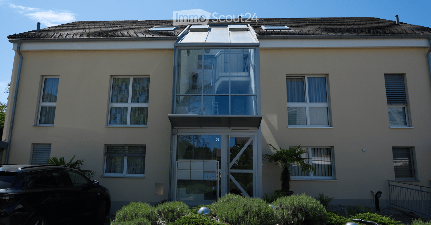 Two-story house with beige exterior, multiple windows, a glass-enclosed balcony, and a small garden area with shrubs and a palm tree.