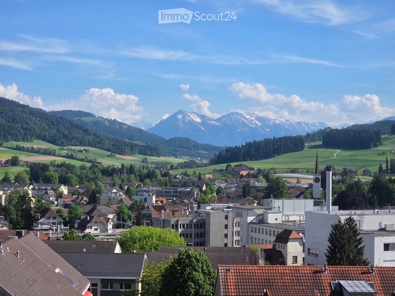 A panoramic view of a town with a mix of residential and commercial buildings, surrounded by green fields and mountains in the background.