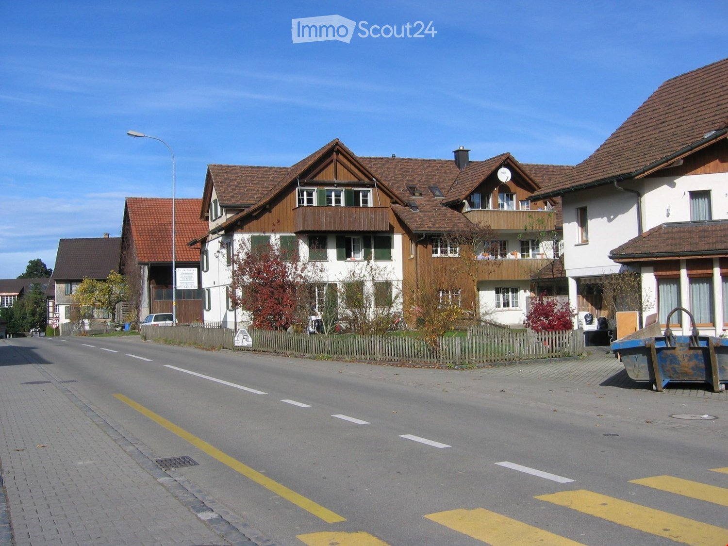 Residential street with traditional Swiss houses featuring wooden facades, tiled roofs, and balconies. The road is paved with clear markings, and there are small fences and trees lining the street.
