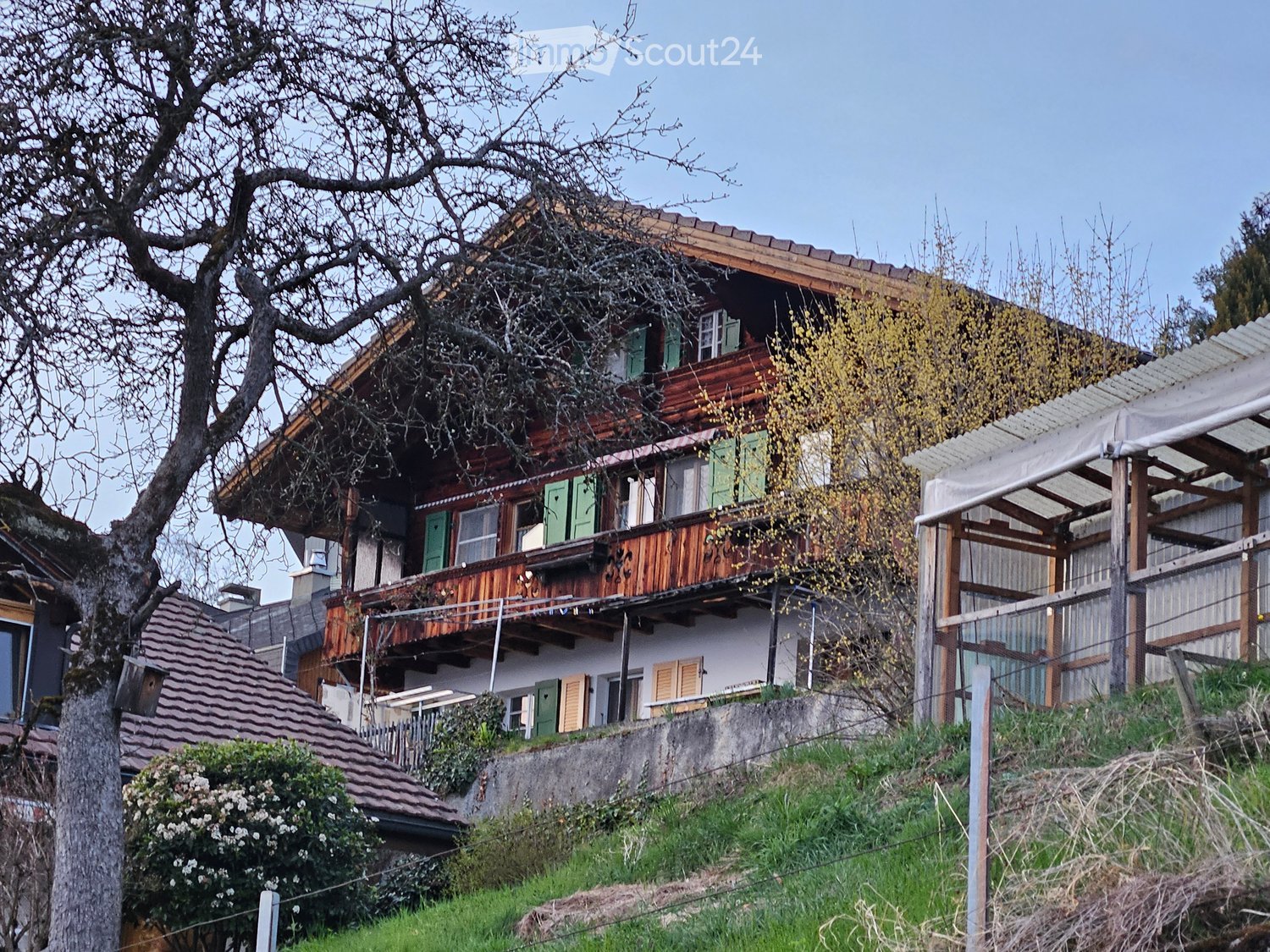 Two-story wooden house with green shutters, wooden balcony, and covered patio area.