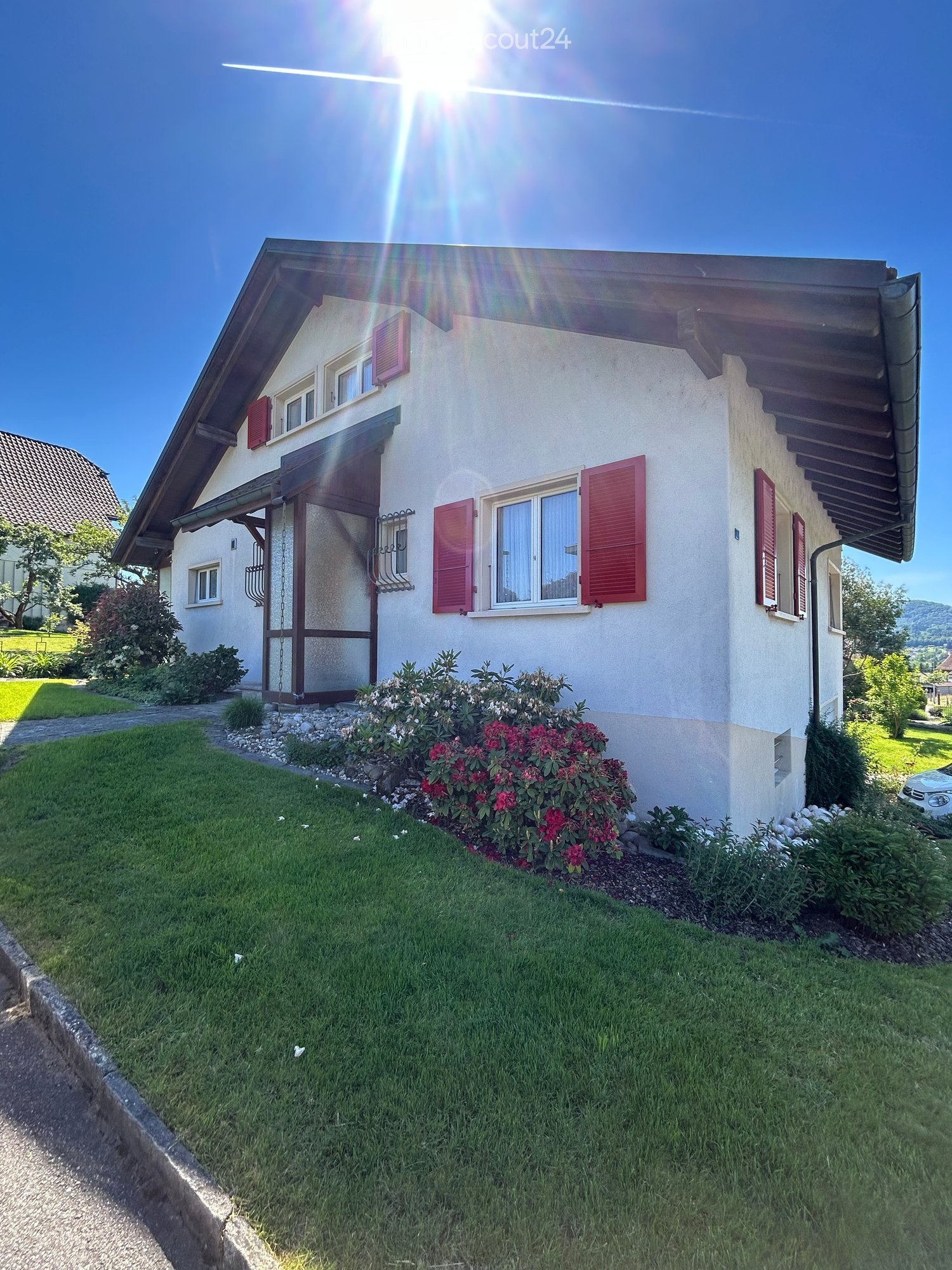 Two-story house with white walls, red shutters, and a well-maintained garden with flowering shrubs and green grass.